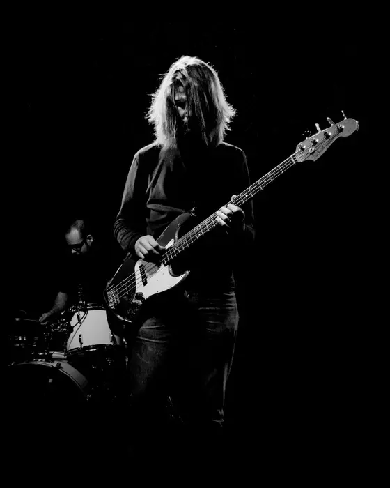A bassist with long blond hair under a single spotlight, black and white.