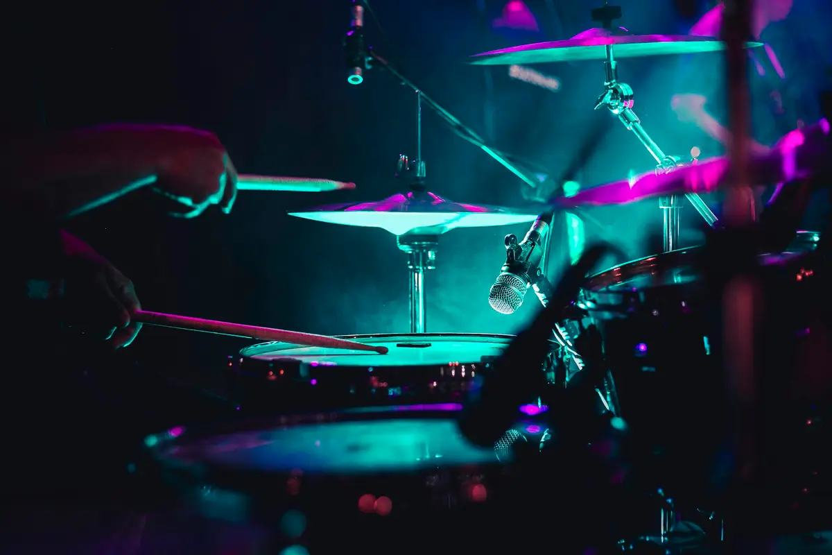 Hands and drumsticks over a kit lit teal and magenta on a dark stage.